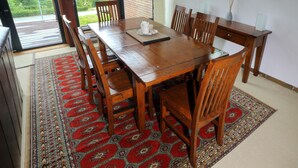 A dining area with a wooden table and chairs, a patterned rug, and a view of greenery through large windows. - Pacific Rim Retreat (Ucluelet)