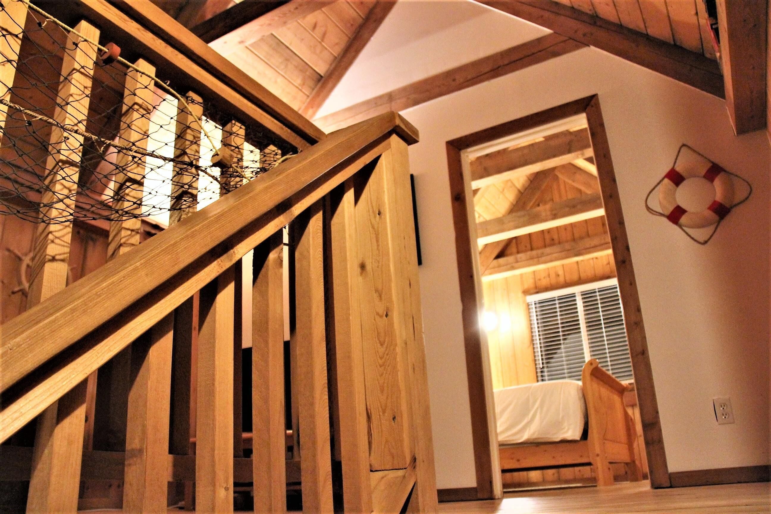 Eine Holztreppe führt zu einem Schlafzimmer auf dem Dachboden, das ein schräges Dach und ein Bett mit weißer Matratze hat.