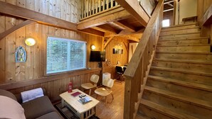 Interior de una cabaña de madera con una escalera, un comedor y una sala de estar con un sofá y una mesa.