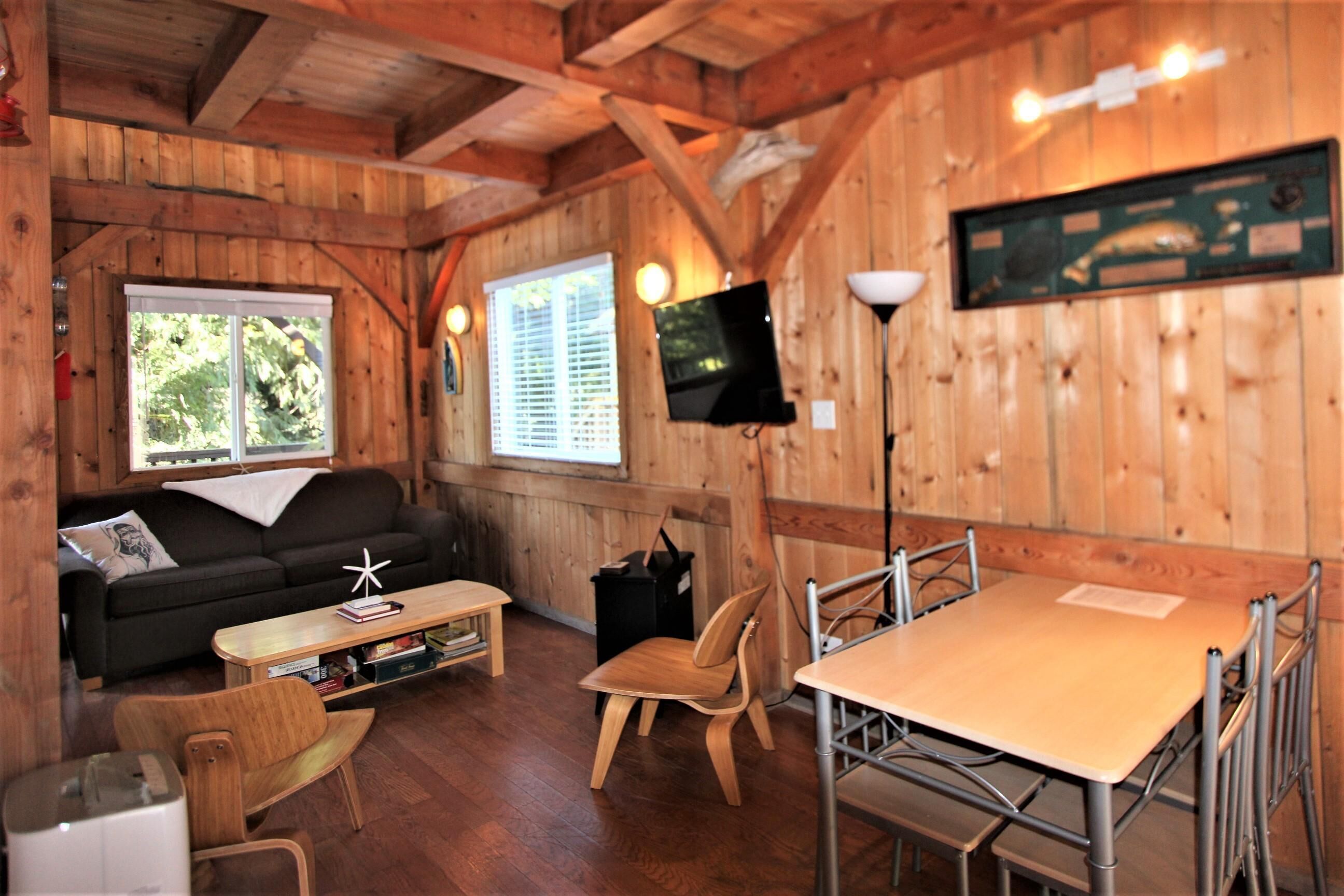 A cozy cabin interior with a sofa, wooden dining table, and a television mounted on the wall.
