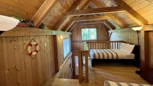Acogedor dormitorio en el ático con vigas de madera, camas literas, una mesita pequeña y ventana con vistas a árboles.