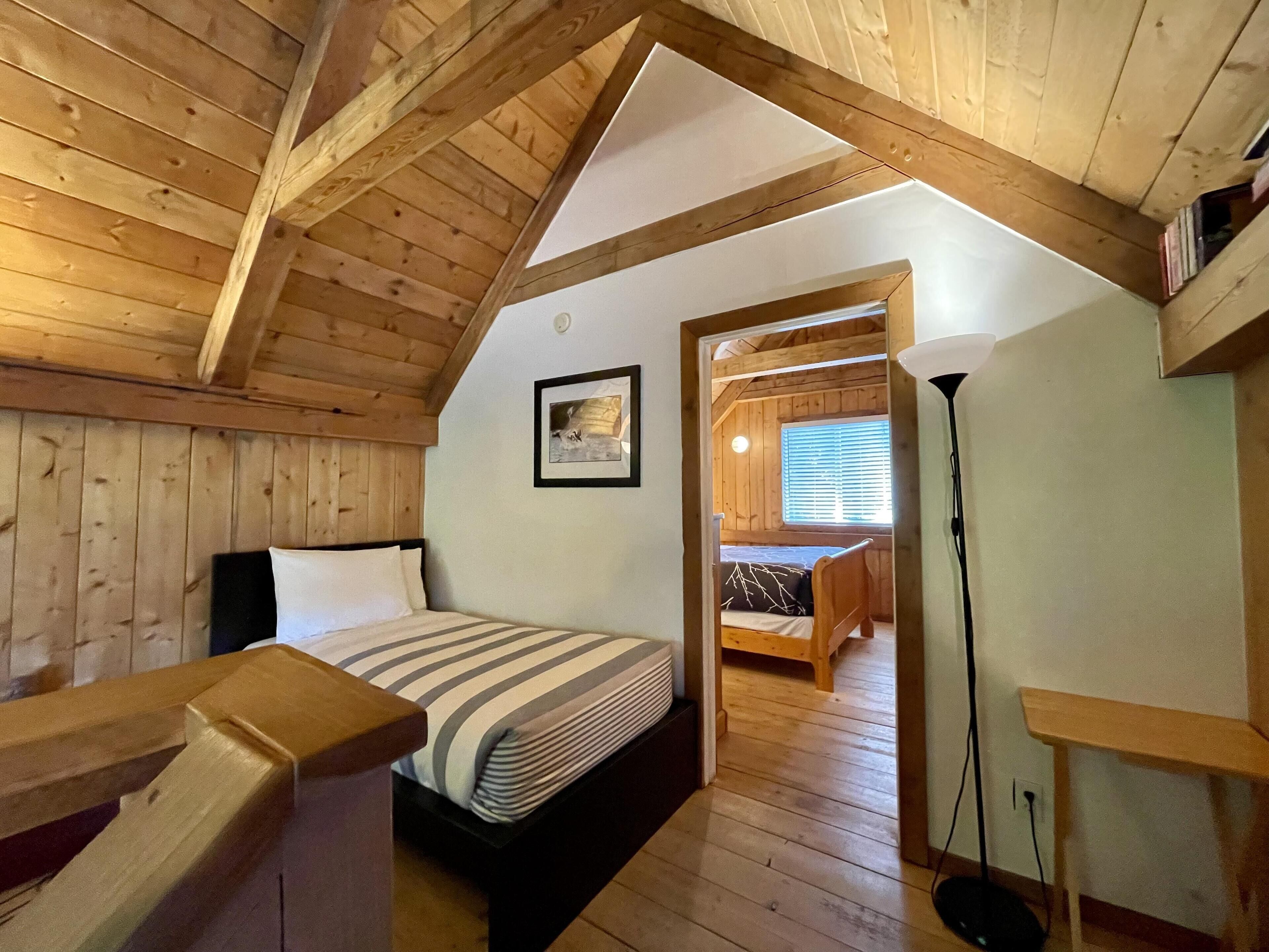 A cozy attic bedroom with a wooden ceiling, a bed with striped bedding, a small desk, and a window with blinds.