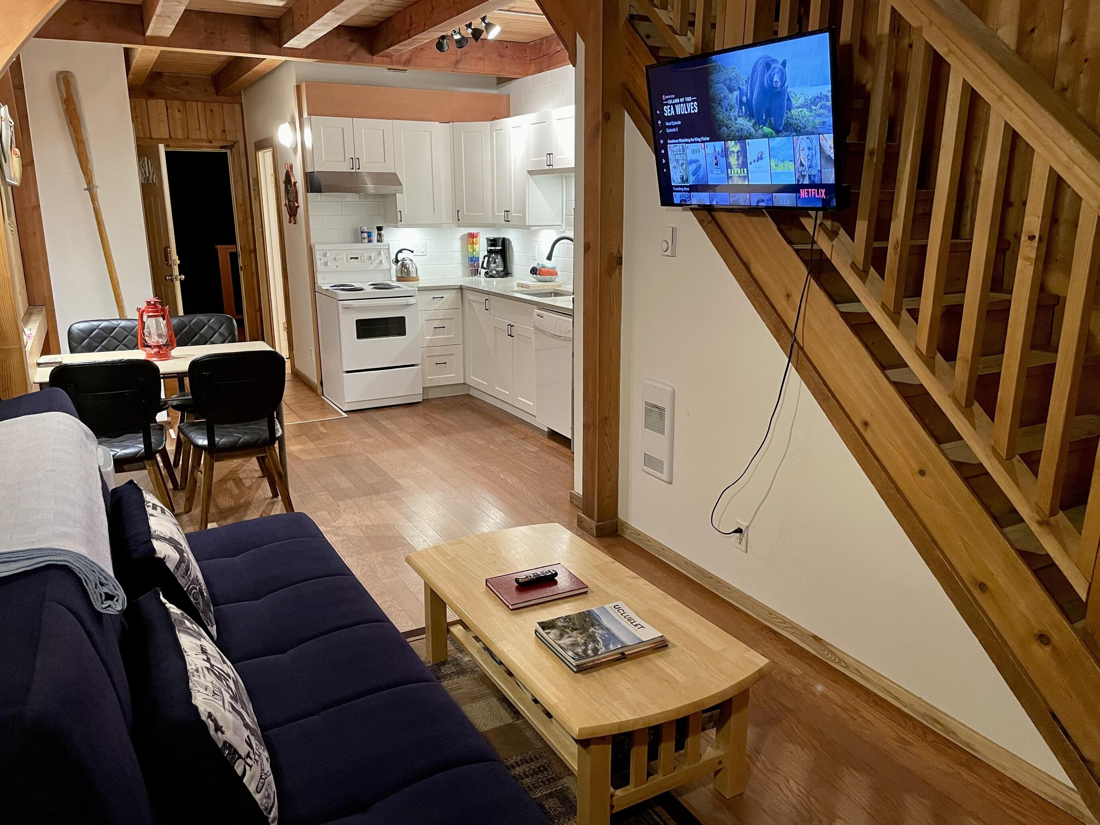 Uma sala de estar aconchegante com um sofá, uma mesa de centro com livros, uma TV fixada em uma parede de madeira e uma área de cozinha com armários brancos e um fogão.
