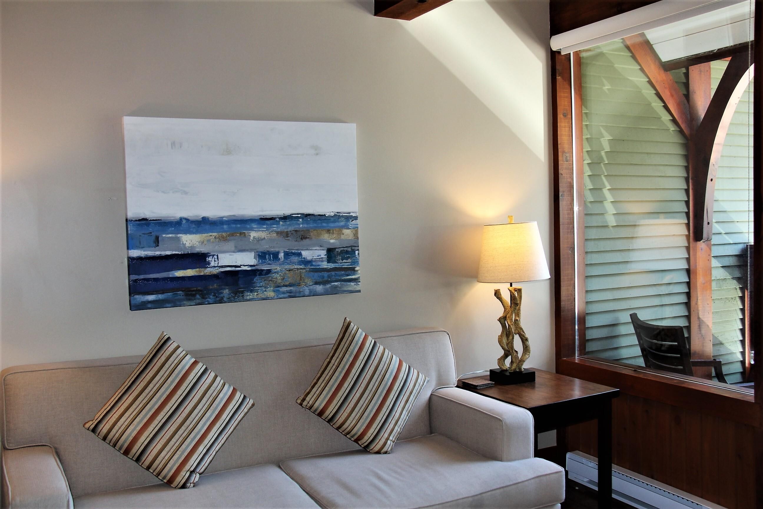 Un salón con sofá, dos almohadas a rayas, una mesa con lámpara y un cuadro en la pared.