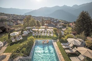 A swimming pool with lounge chairs and umbrellas, surrounded by a residential area with houses and mountains in the background.