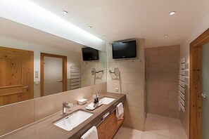 A modern bathroom with two sinks, a large mirror, a television mounted above the mirrors, and a walk-in shower.