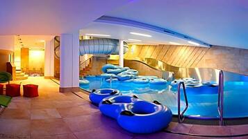 An indoor swimming pool with multiple lanes, a central slide, and seating areas with red cushions.