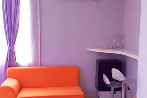 Een kamer met een paarse wand, een witte plank met glazen, een zwarte kast en een oranje bank.