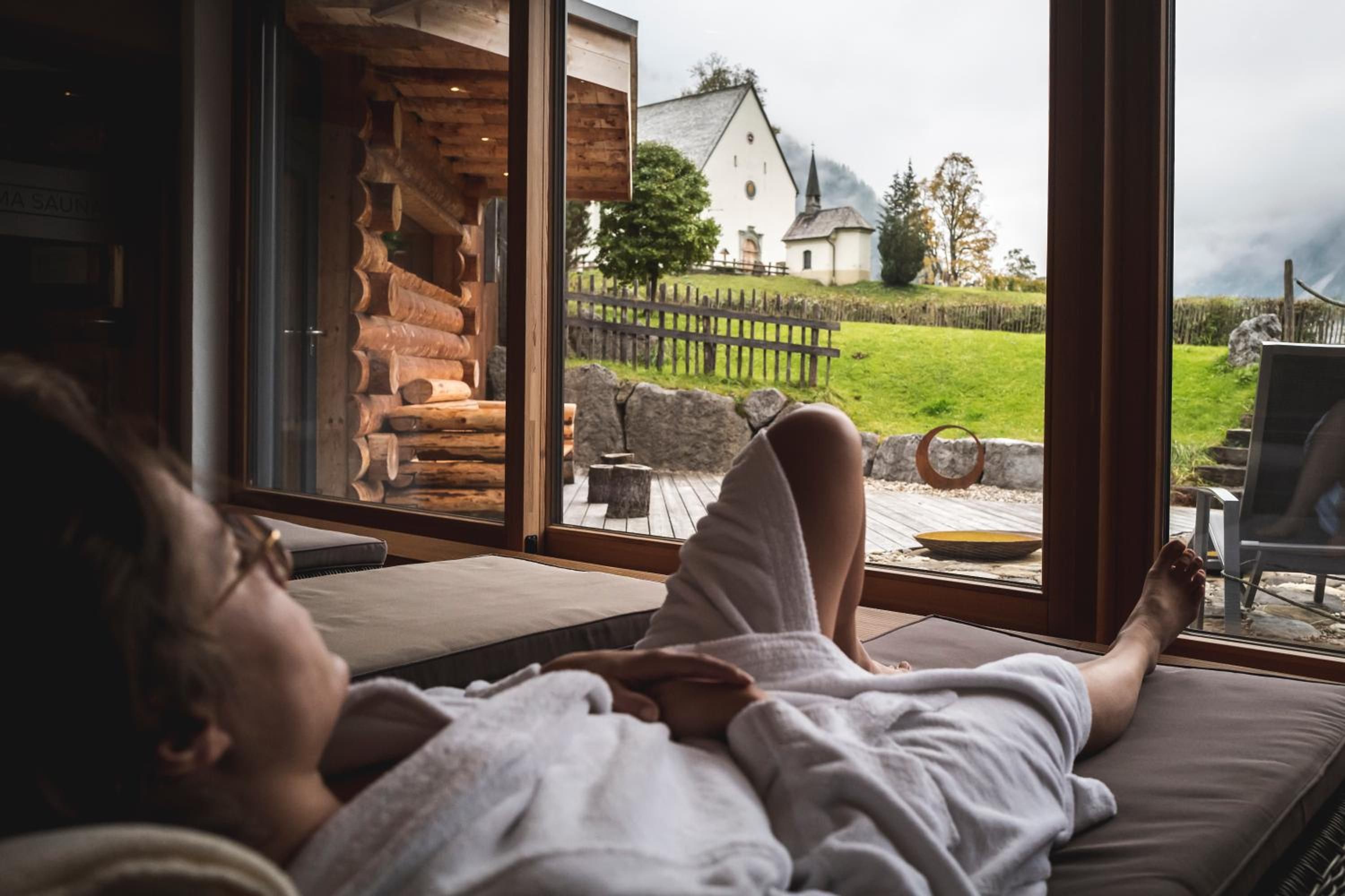 Ein Zimmer mit großem Fenster, das auf eine Holzhütte und ein weißes Gebäude mit Kirchturm blickt.