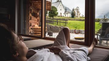 Ein Zimmer mit großem Fenster, das auf eine Holzhütte und ein weißes Gebäude mit Kirchturm blickt.