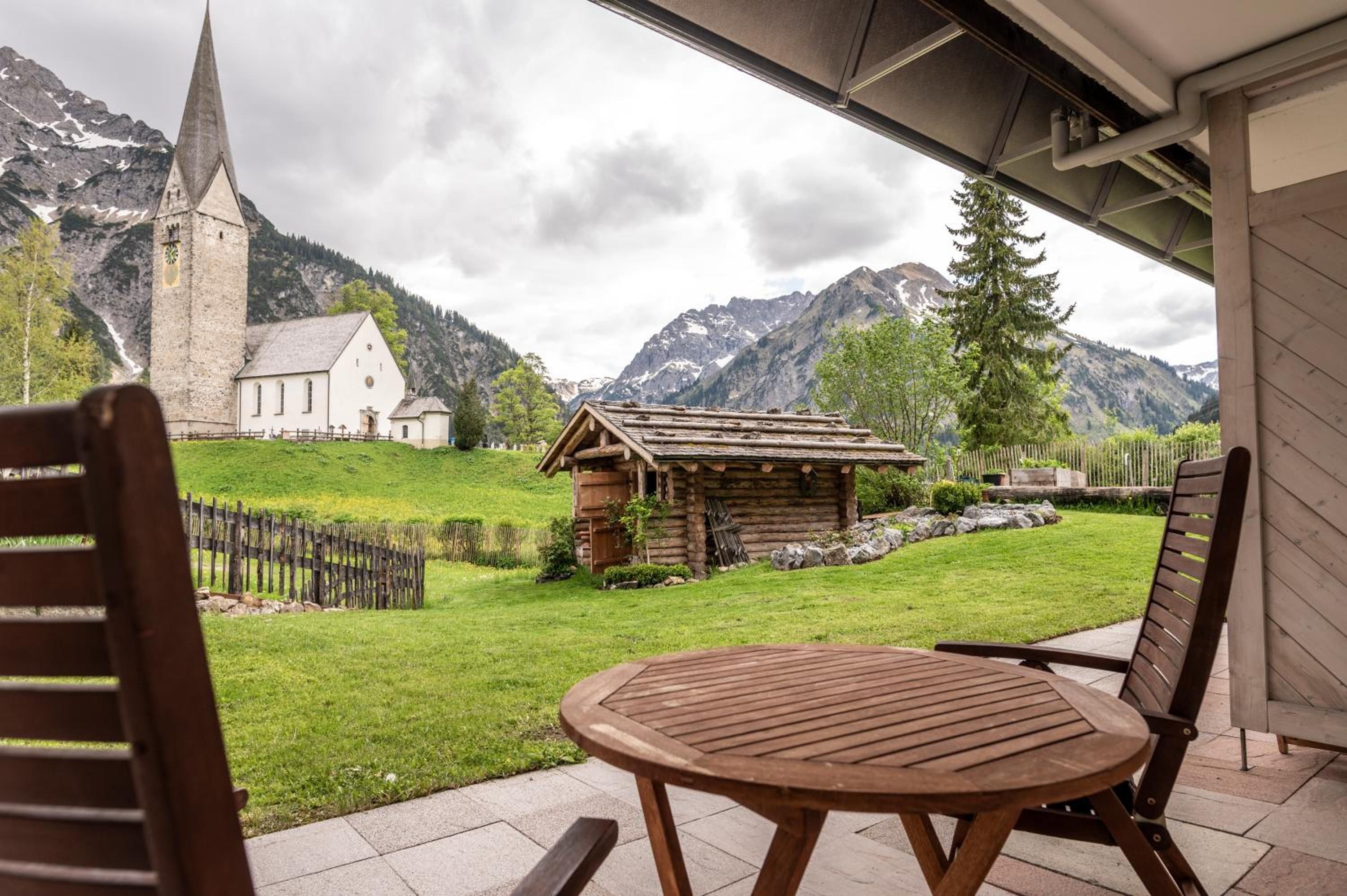 Ein Holztisch mit Stühlen auf einer Terrasse, mit Blick auf eine Kirche und Berge.