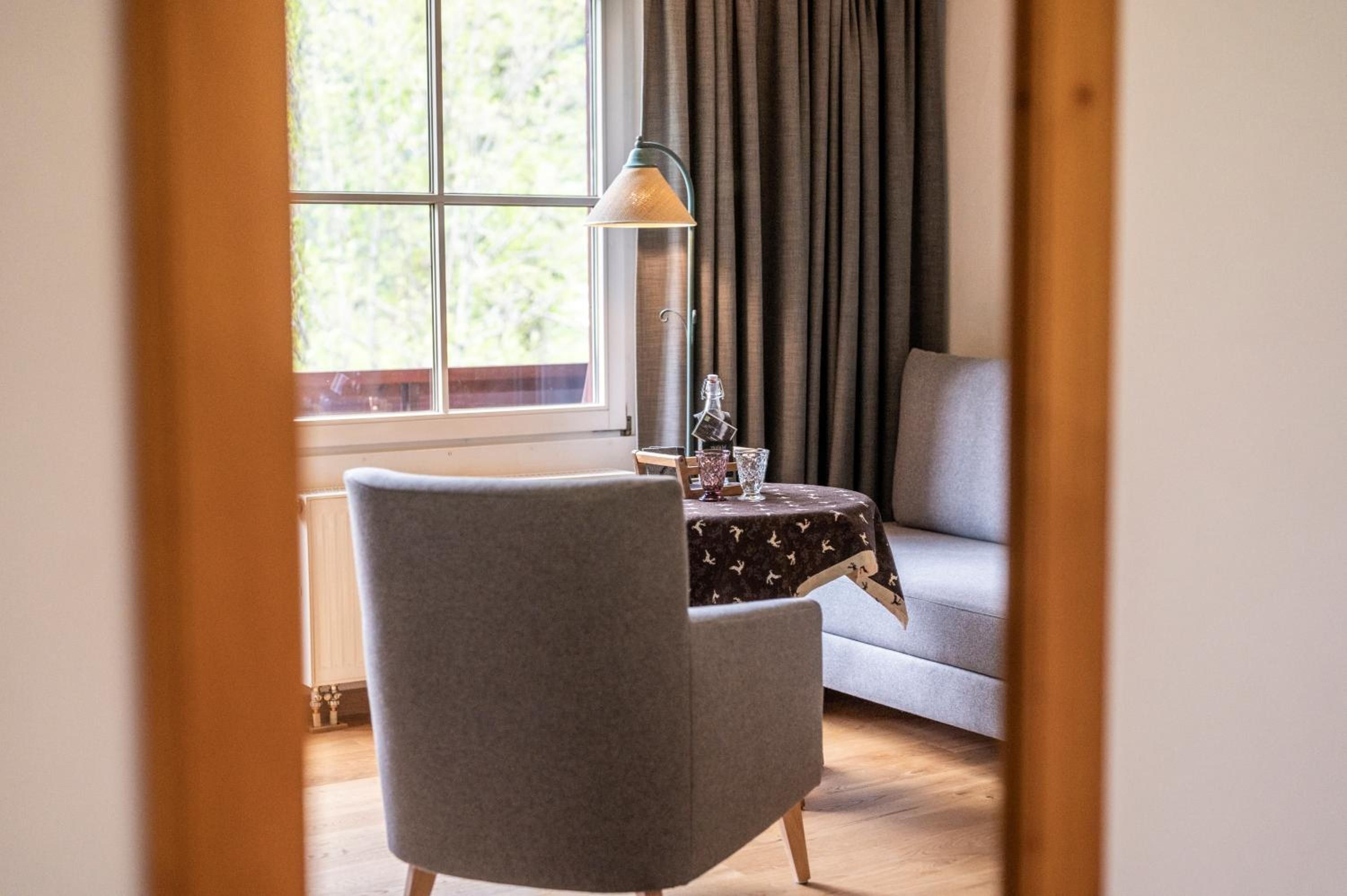 A room with a grey armchair, a small table with a patterned tablecloth, a lamp, and a window with curtains.