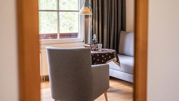 A room with a grey armchair, a small table with a patterned tablecloth, a lamp, and a window with curtains.