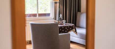 A room with a grey armchair, a small table with a patterned tablecloth, a lamp, and a window with curtains.