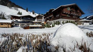 Schneebedeckte Landschaft mit einem großen Holzhäuschen mit mehreren Balkonen und einer Seilbahn im Hintergrund.
