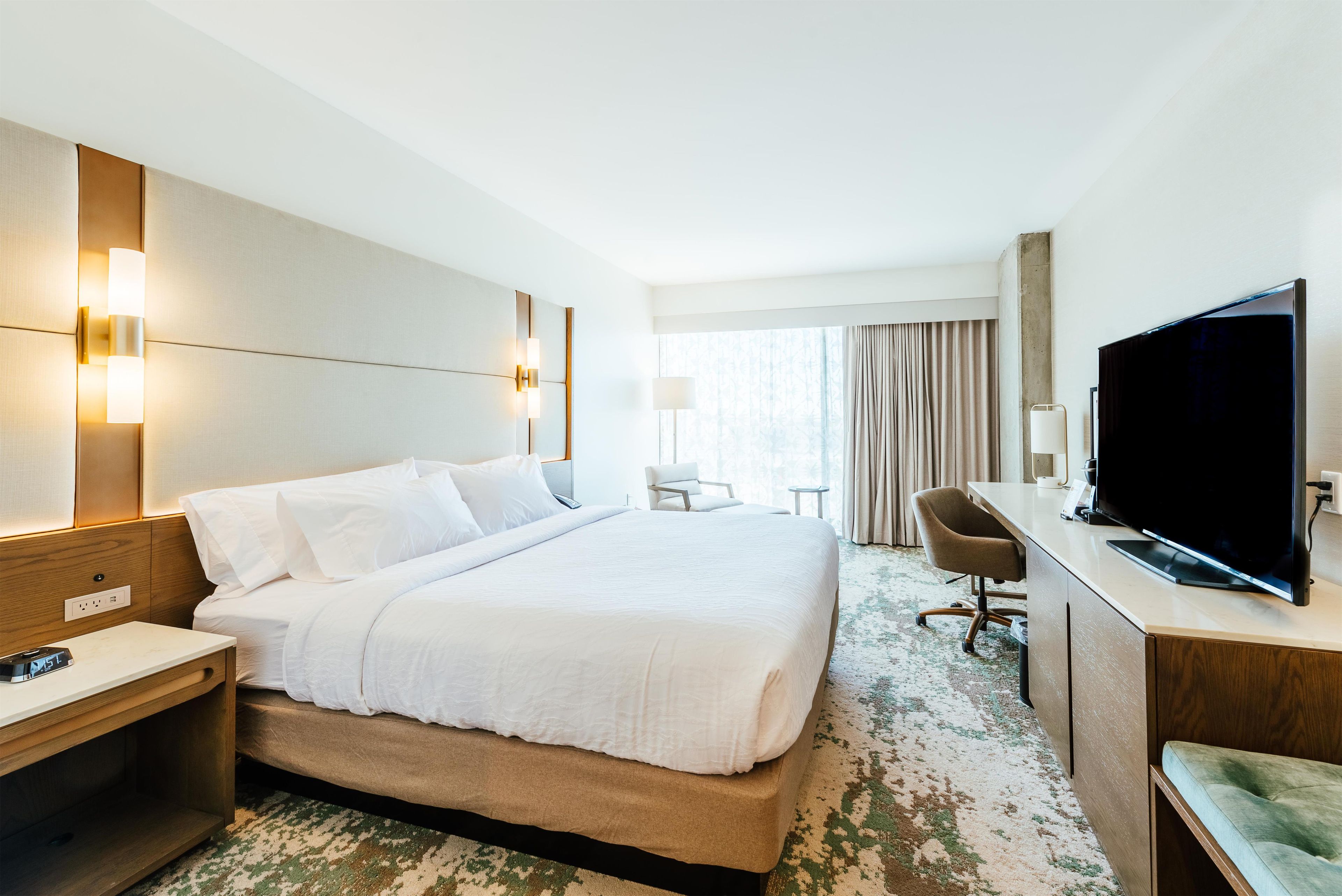 Ein Hotelzimmer mit einem großen Bett, Nachttischen, einem Schreibtisch mit Flachbildfernseher, einem Sessel und einem Fenster mit Vorhängen.