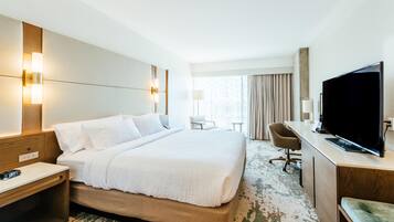Ein Hotelzimmer mit einem großen Bett, Nachttischen, einem Schreibtisch mit Flachbildfernseher, einem Sessel und einem Fenster mit Vorhängen.