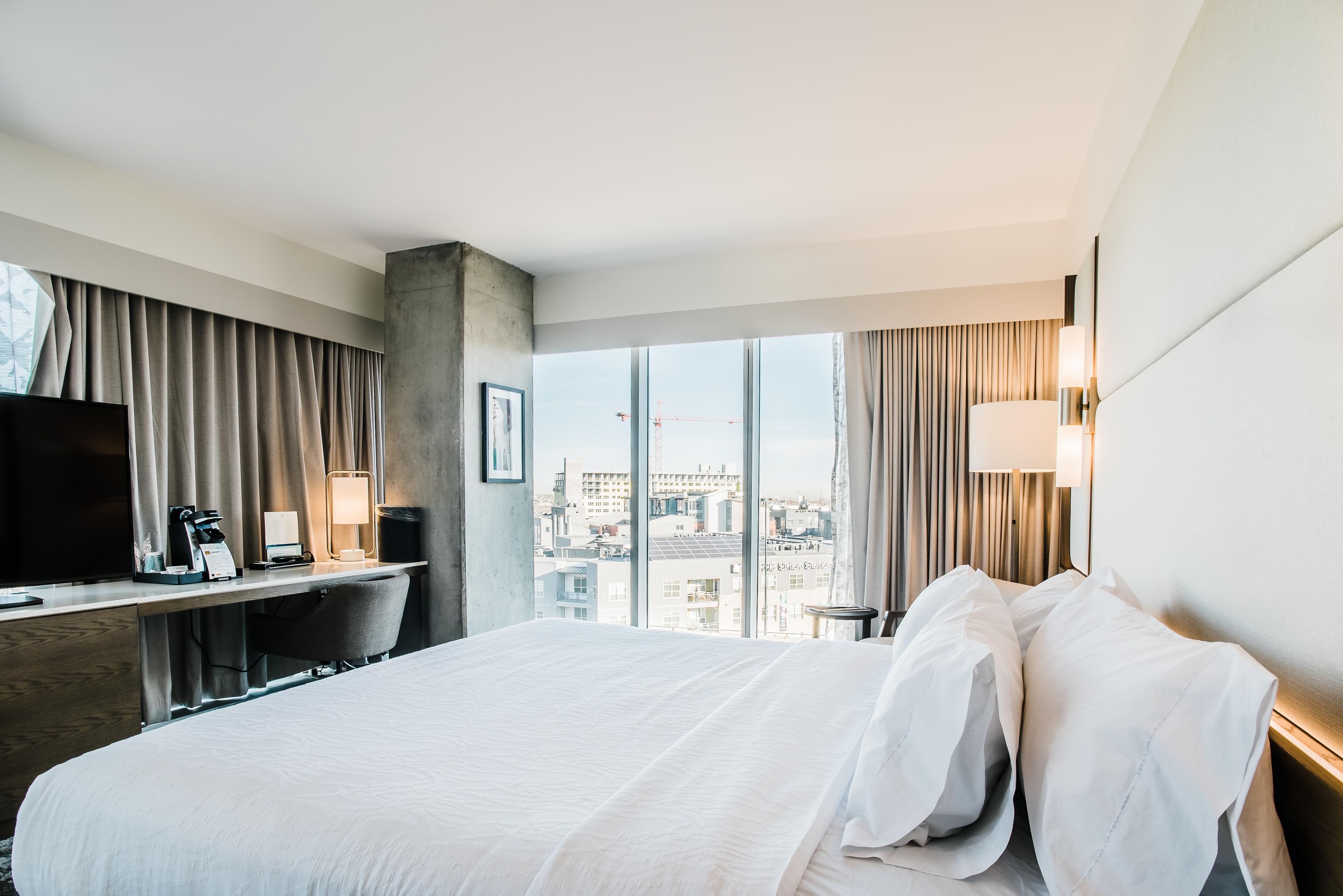 A hotel room with a large bed, a desk with a computer, a TV, and a view of the cityscape.