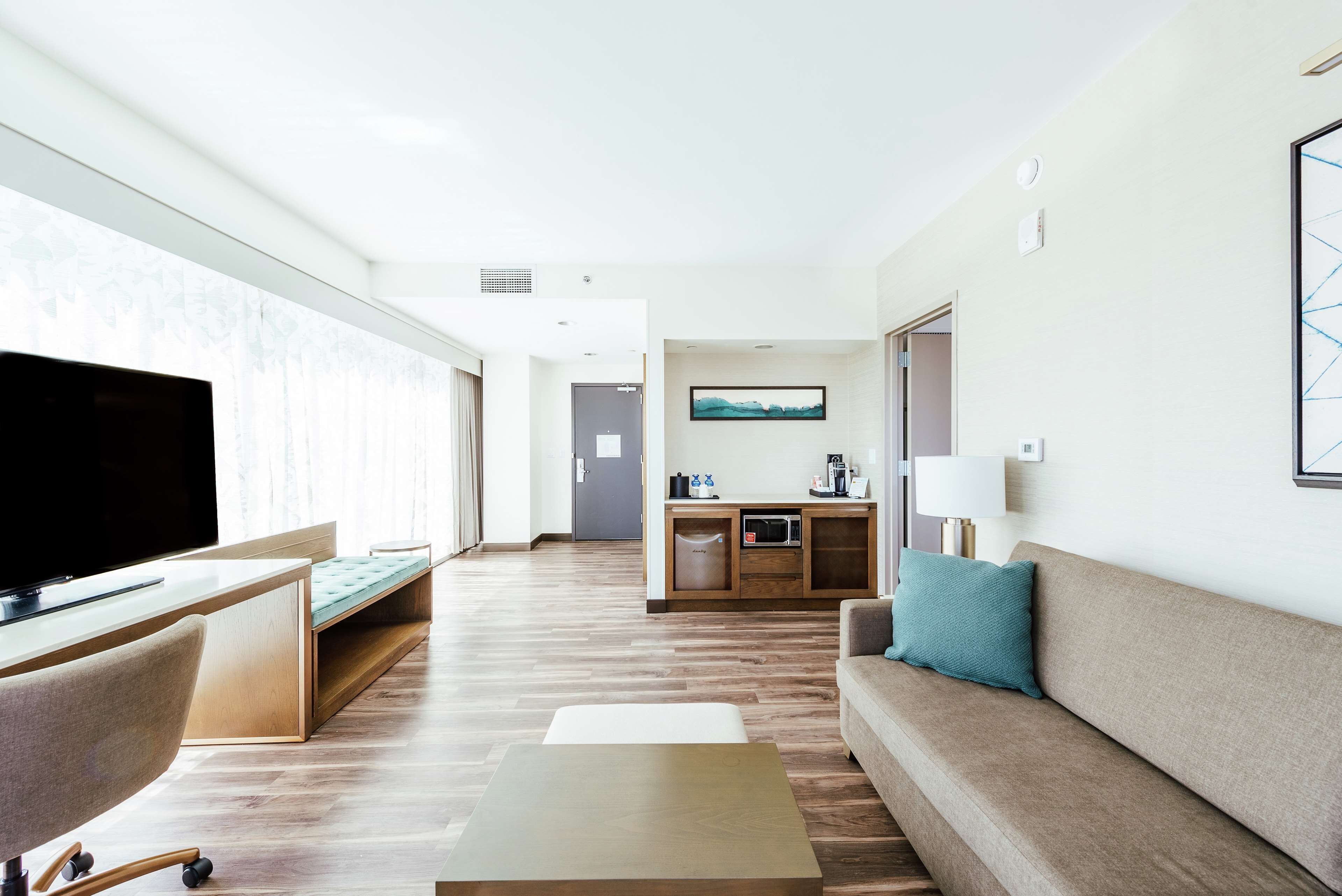 A modern hotel room with a flat-screen TV, a sofa, a coffee table, and a kitchenette area.