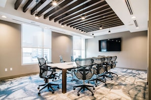 A conference room with a large table, multiple office chairs, a flat-screen TV on the wall, and large windows allowing natural light.