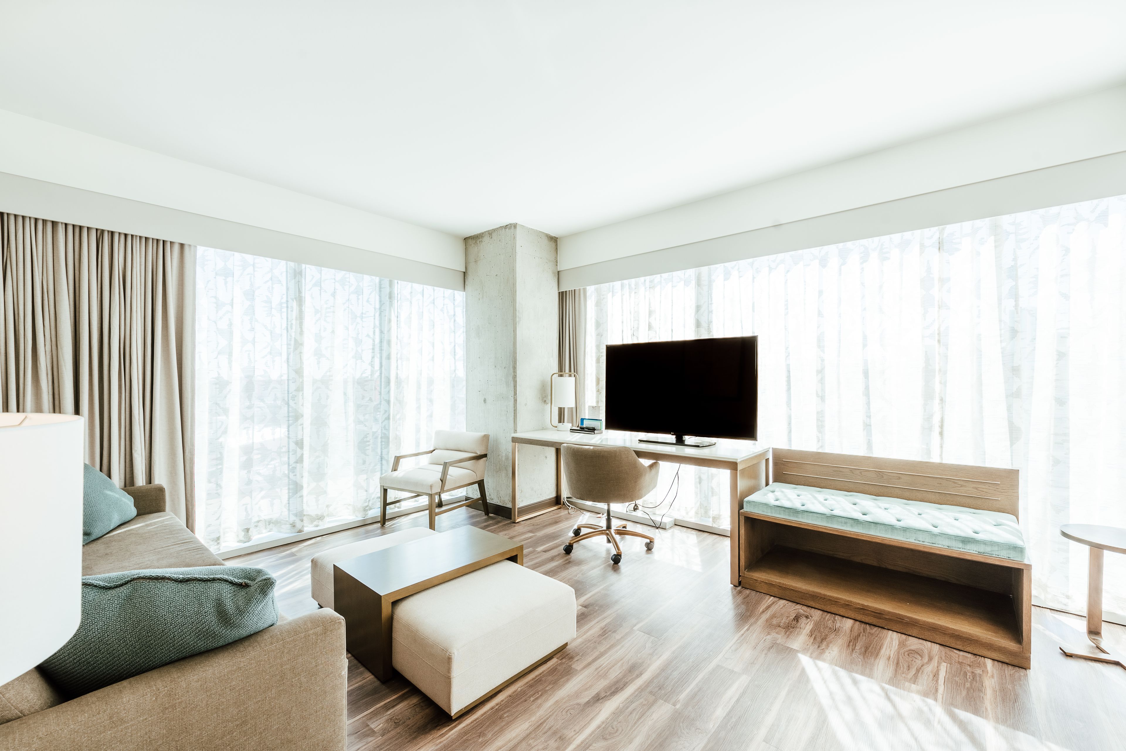 A modern hotel room with a sofa, a desk with a computer, a television, and a large window with curtains.