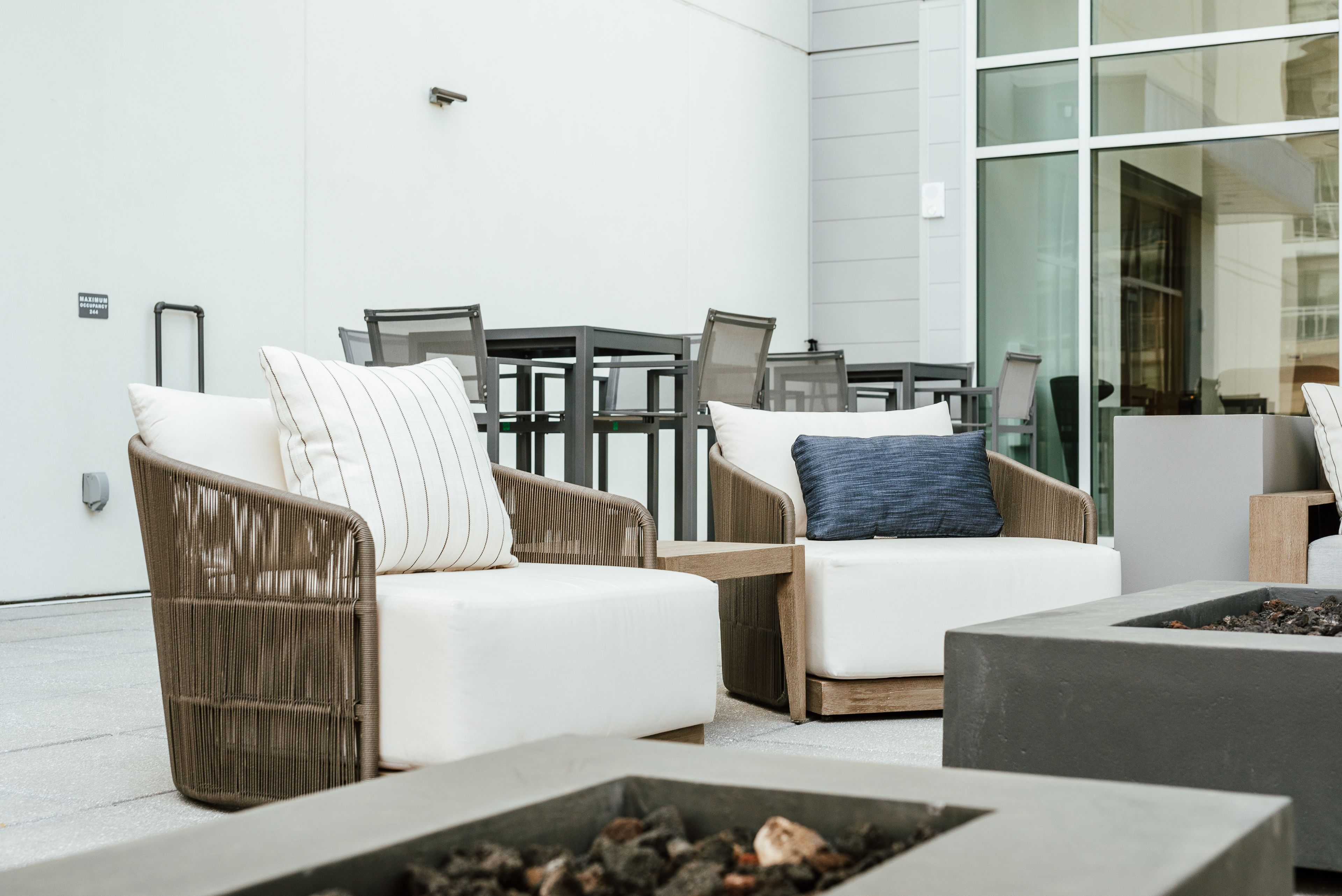 A modern outdoor seating area with wicker chairs and sofas, a fire pit, and a glass wall.