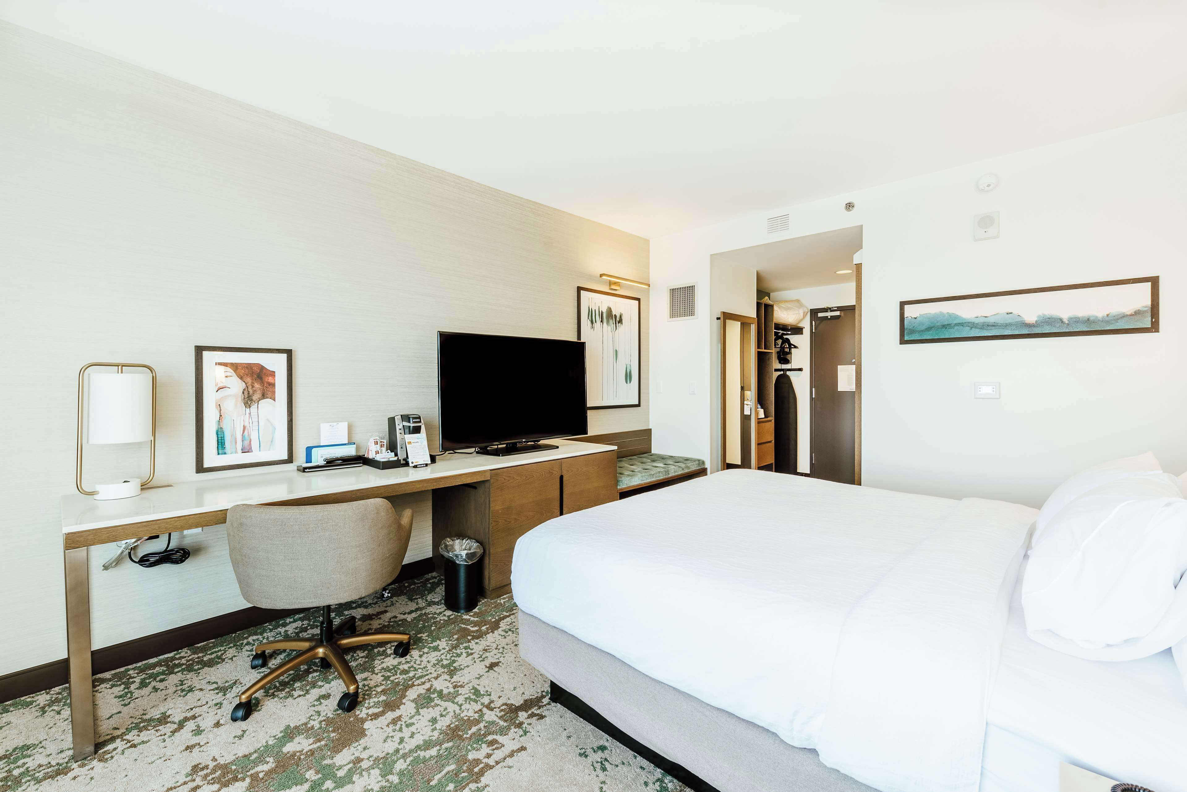 A hotel room with a bed, desk, chair, television, and artwork.