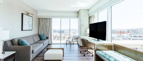 A modern living room with a sofa, a coffee table, a desk with a chair, a television, and large windows offering a city view.