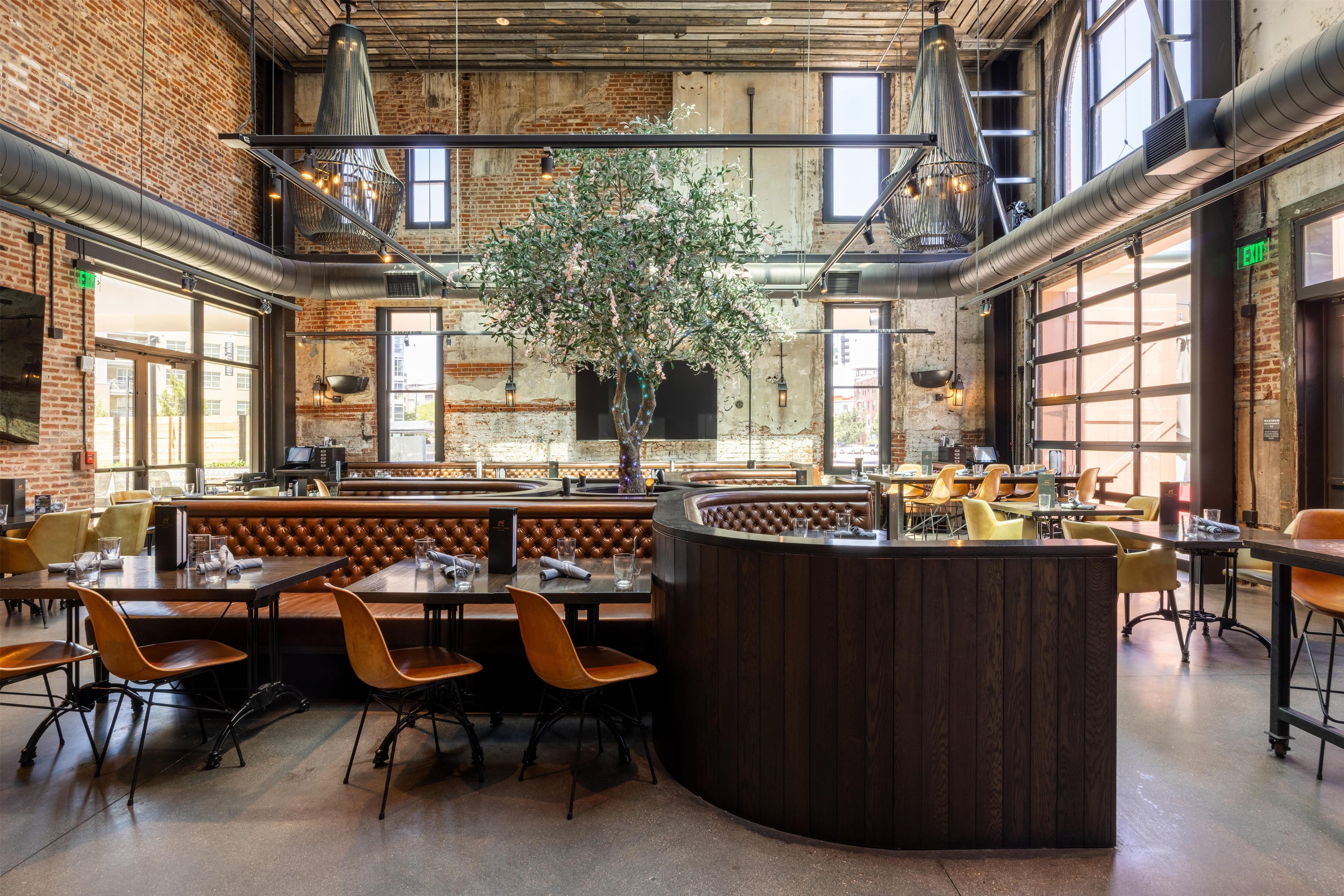 A restaurant with brick walls, large windows, and a central plant. Tables and chairs are arranged for dining.