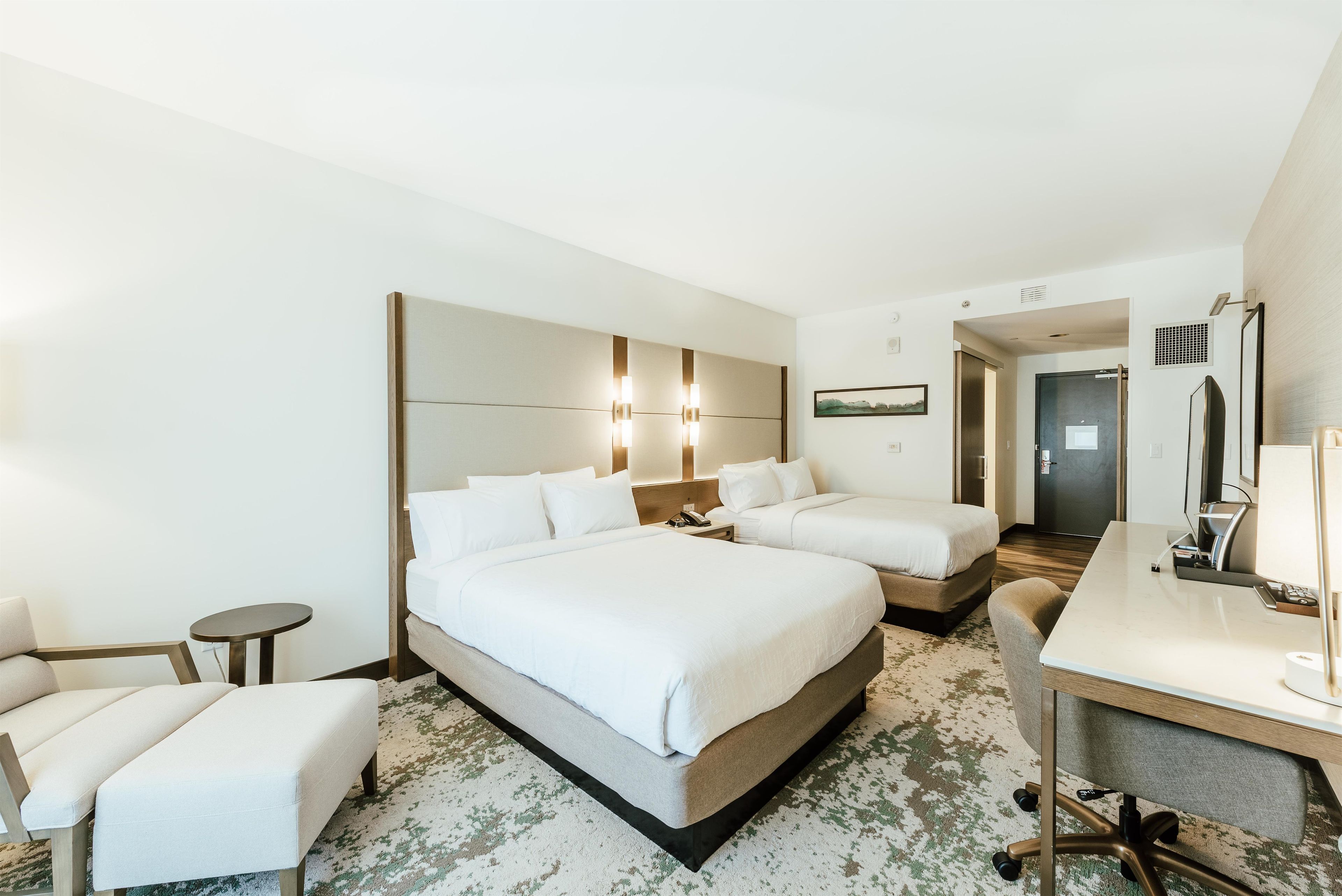 A hotel room with two beds, a desk, a chair, a small round table, and a bathroom visible through an open door.