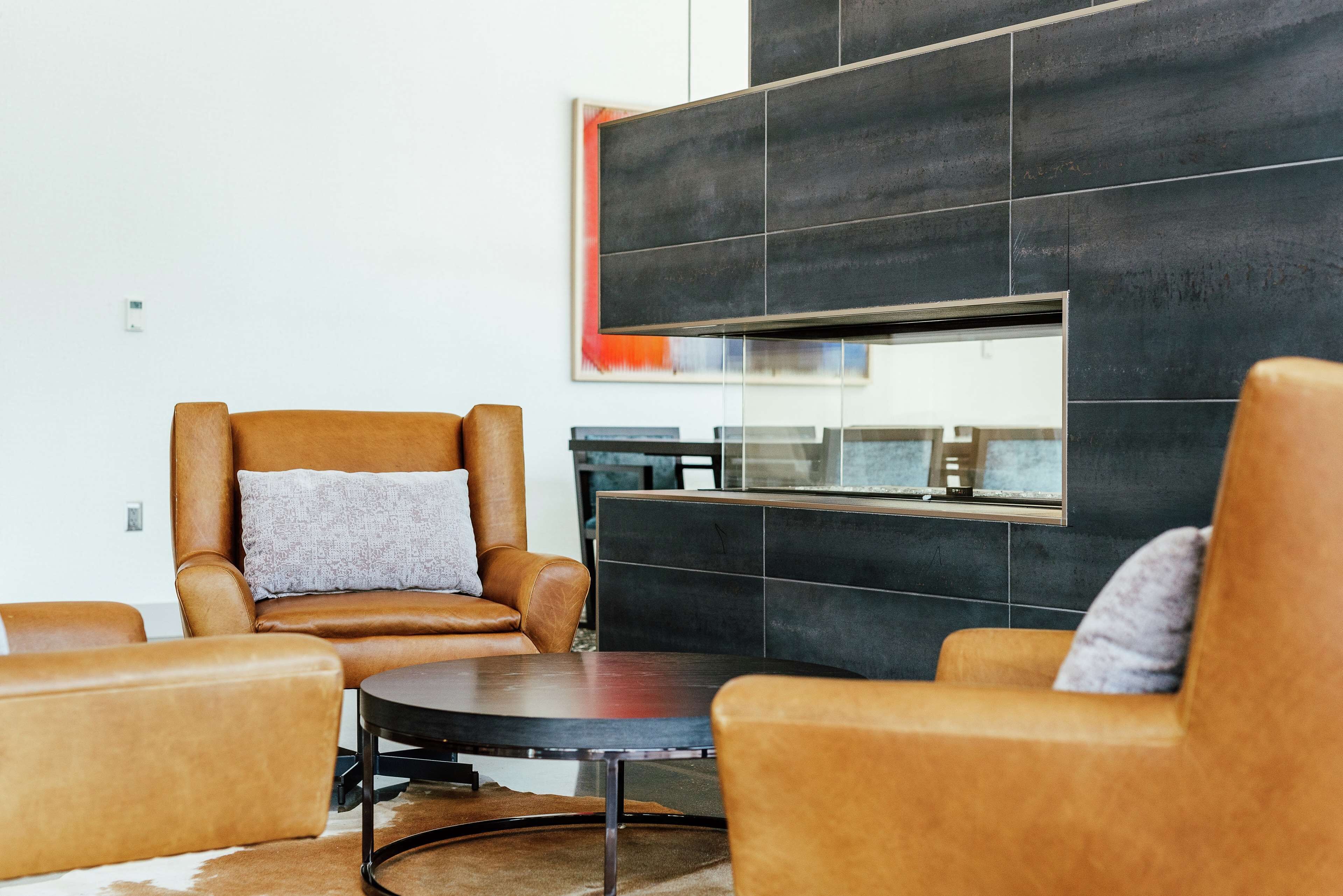 A modern living room with leather armchairs, a round coffee table, and a large black-tiled wall with a framed artwork.