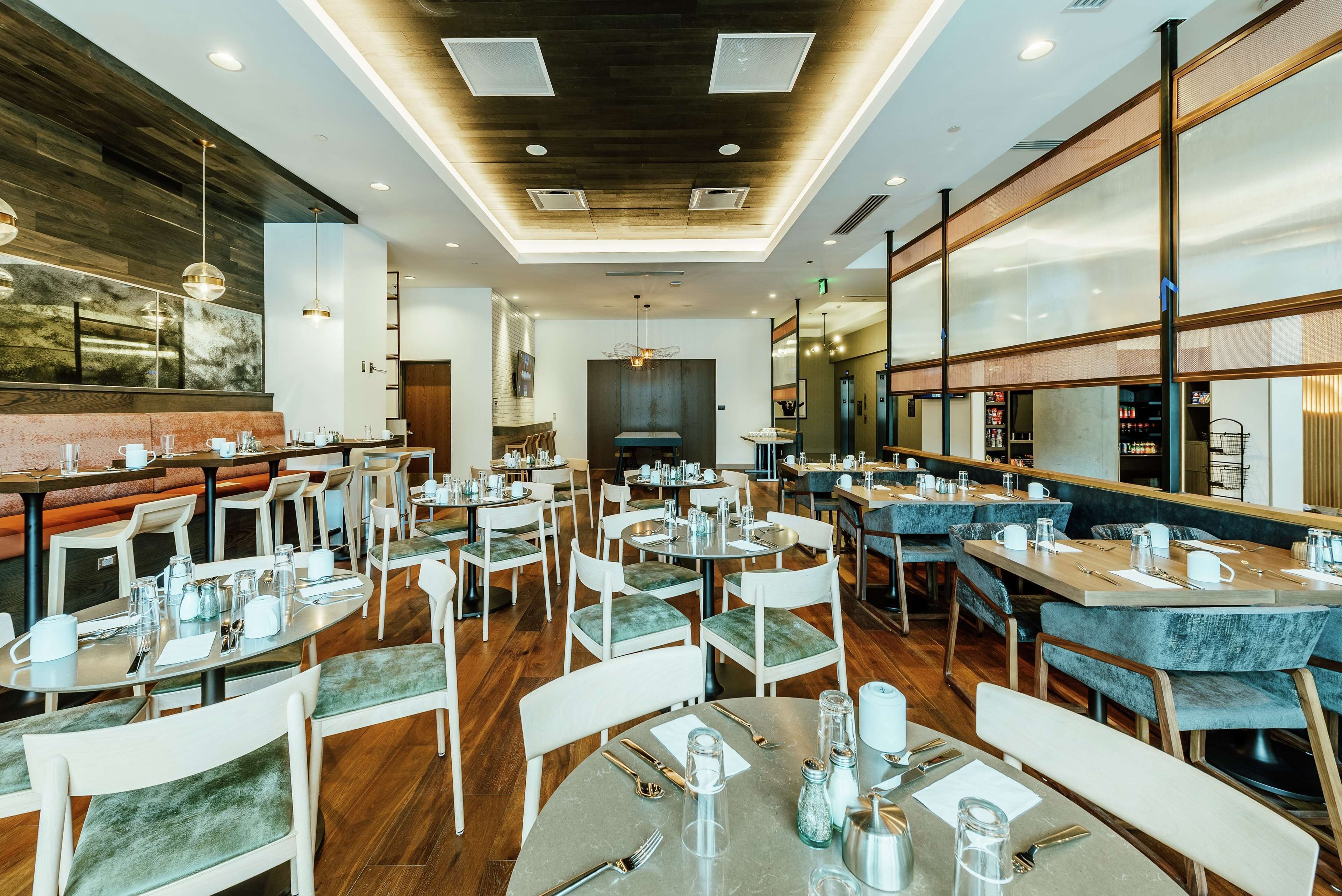 A modern restaurant with round tables and booths, wooden flooring, and a high ceiling with recessed lighting.