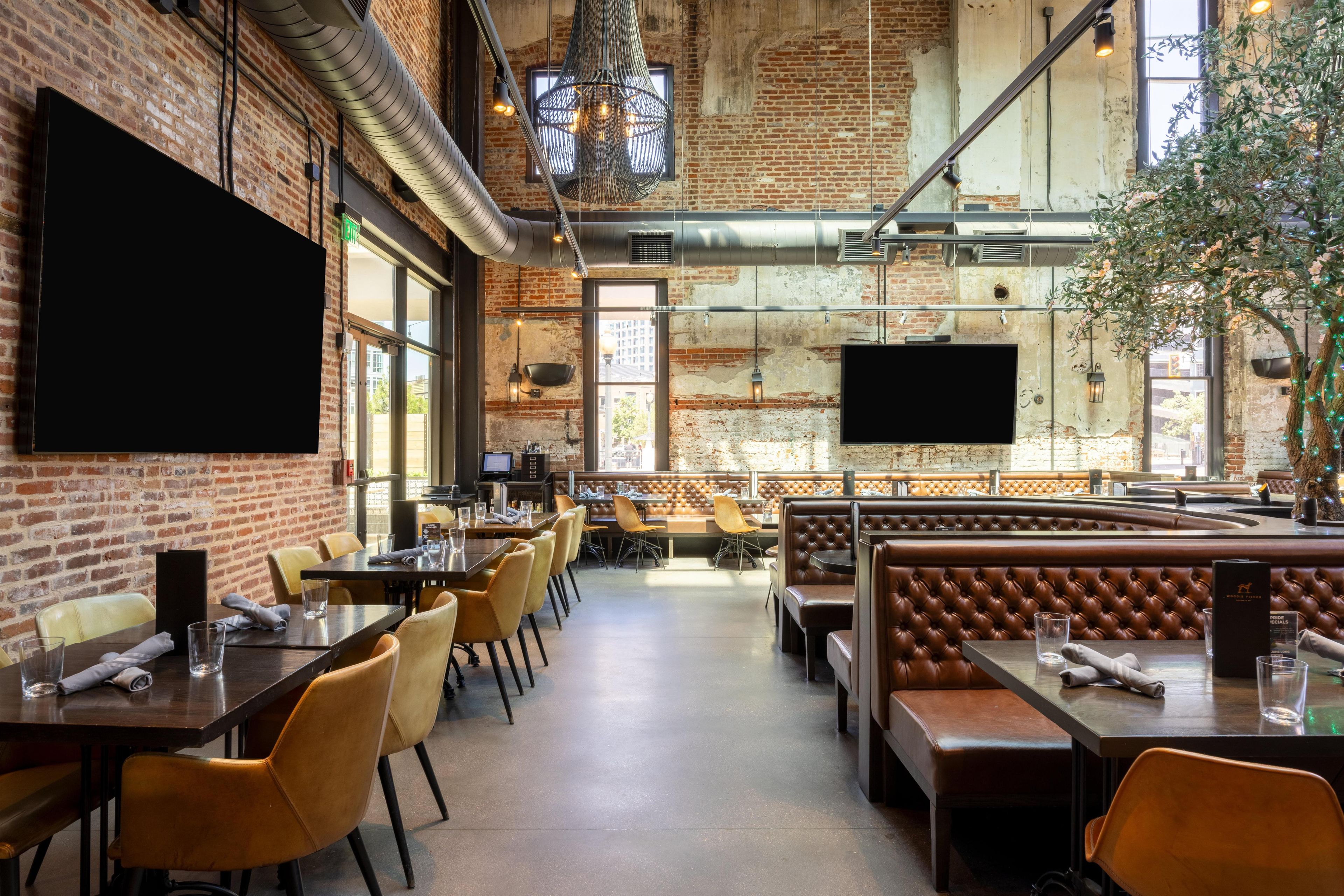 A restaurant with brick walls, large windows, and a high ceiling. Tables and chairs are arranged for dining.