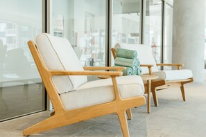 Ein moderner Lounge-Bereich mit Holzstühlen und einem kleinen runden Tisch, großen Fenstern mit Blick auf Gebäude sowie einer Glaswand.