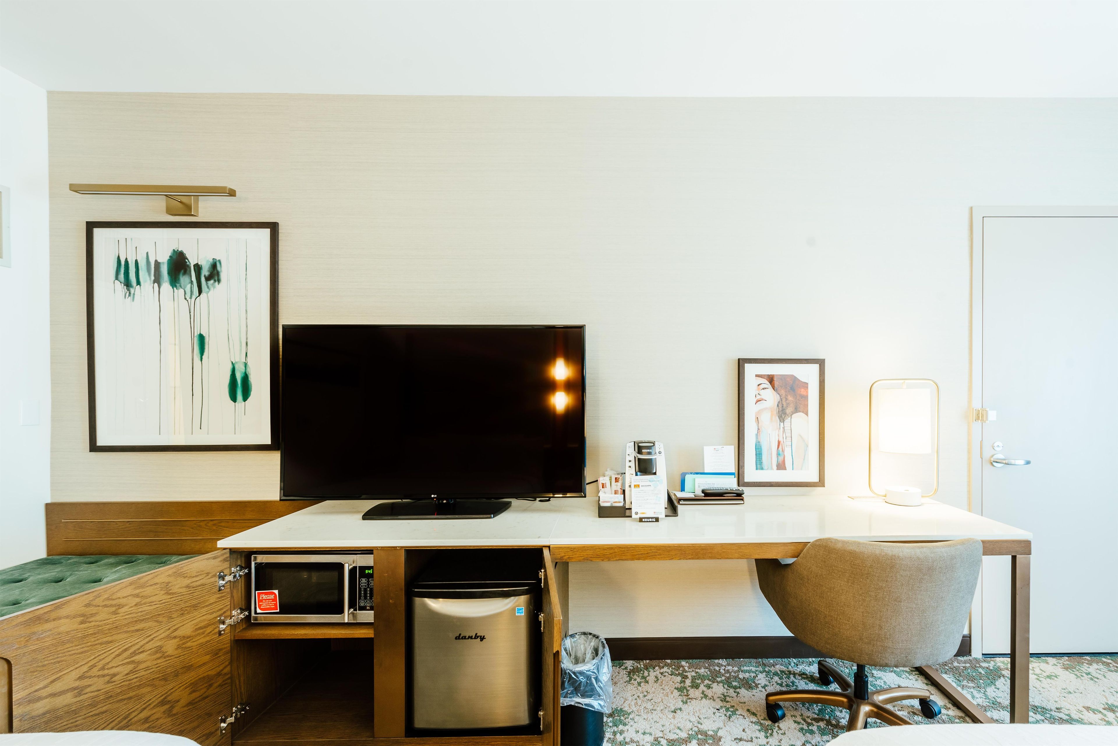 Habitación de hotel con televisión de pantalla plana, minibar, escritorio con lámpara y un cuadro en la pared.