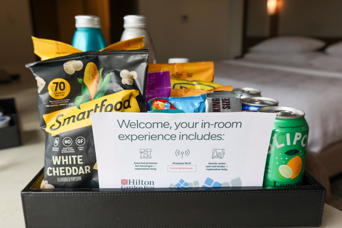 Ein Hotelzimmer mit einer Willkommensanzeige, die Annehmlichkeiten wie Smartfood-Snacks, einer Dose LIPO und ein Schild für Hilton Garden Inn beschreibt.