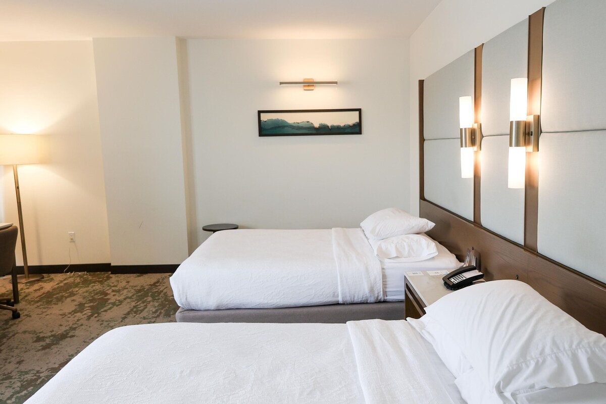 Ein Hotelzimmer mit zwei Betten, einem Nachttisch, einer Lampe, einem Bild an der Wand und einem Telefon auf dem Nachttisch.