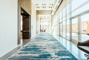 Un pasillo de hotel con alfombra azul estampada, ventanas amplias y una zona de estar a la derecha.