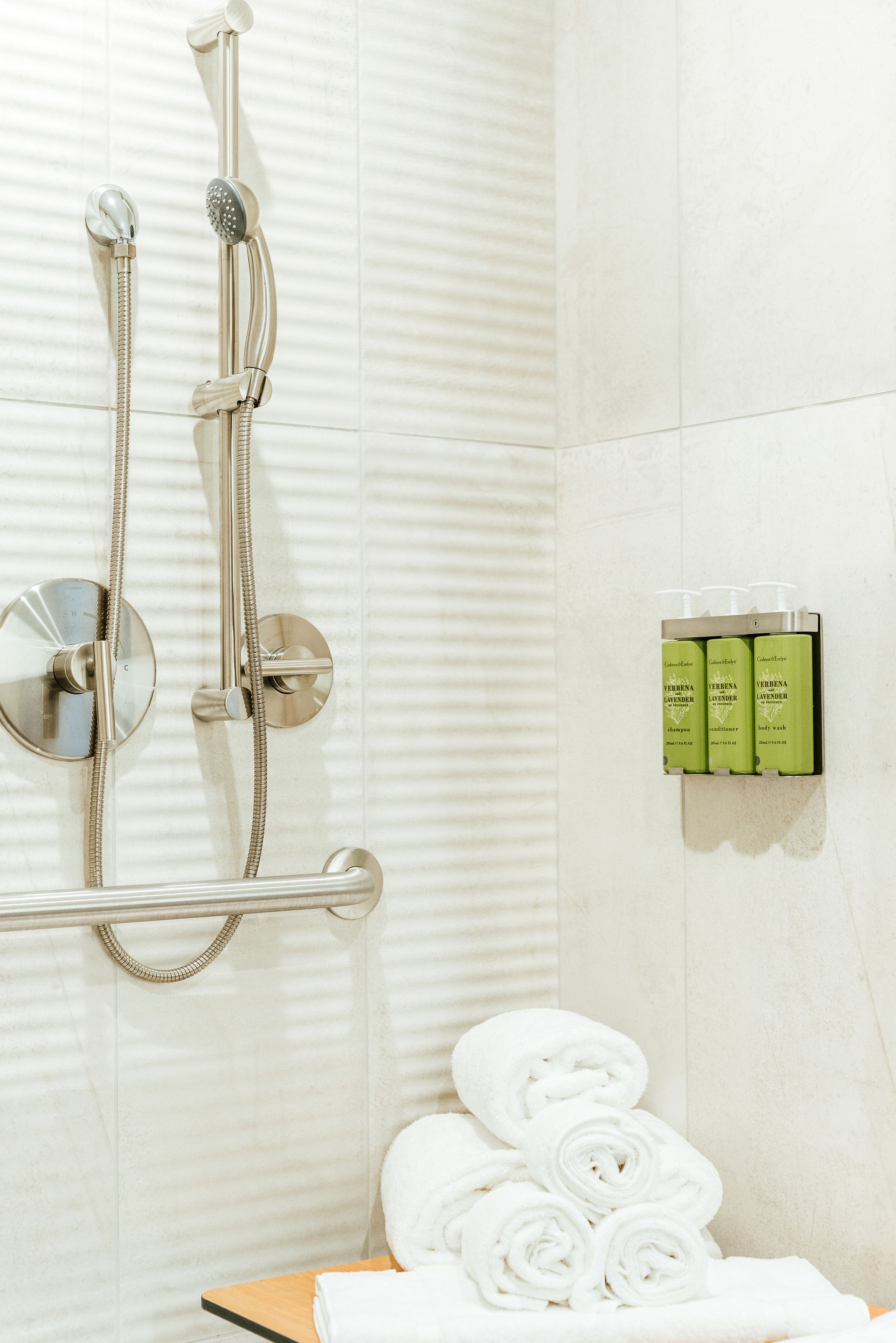 A shower with a handheld showerhead and a wall-mounted faucet, a towel rack with folded towels, and a set of toiletries on the wall.