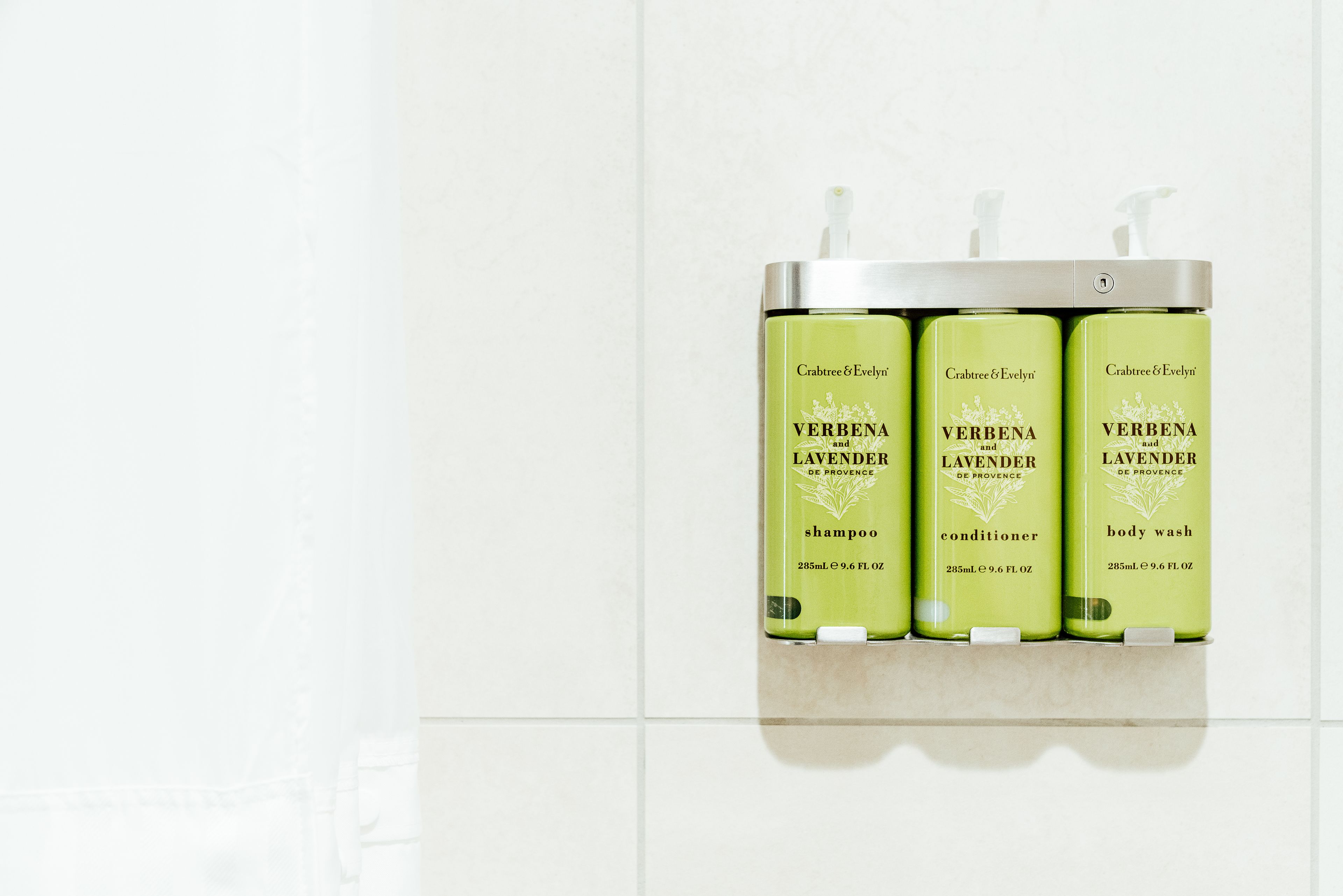 Three bottles of Crabtree & Evelyn Verbena Lavender De Provence shampoo, conditioner, and body wash on a bathroom shelf.