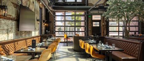 A restaurant with high ceilings, exposed brick walls, and large windows. Tables and chairs are arranged for dining.