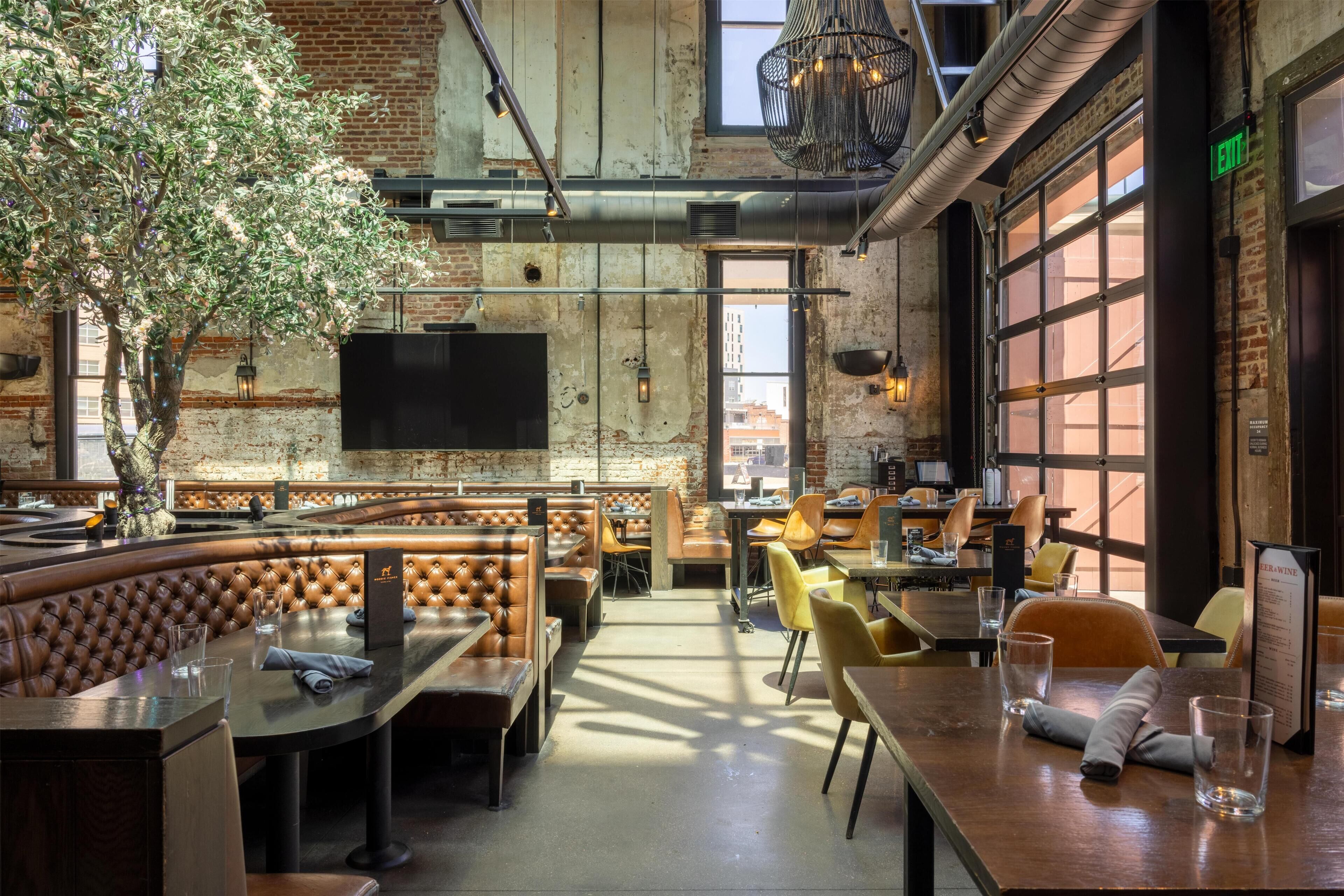 Restaurante com pé direito alto, paredes de tijolo aparente e uma variedade de assentos, incluindo cabines e mesas.