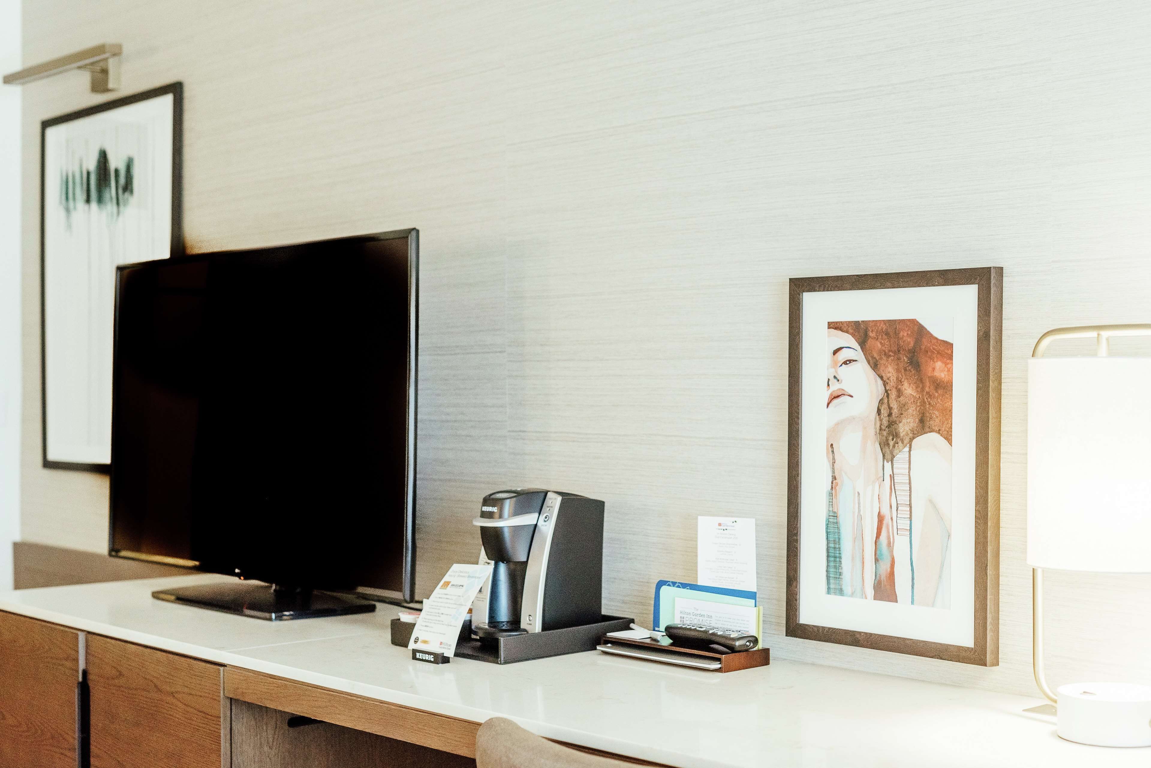 A hotel reception desk with a flat-screen TV, a coffee machine, a framed artwork, and a lamp.