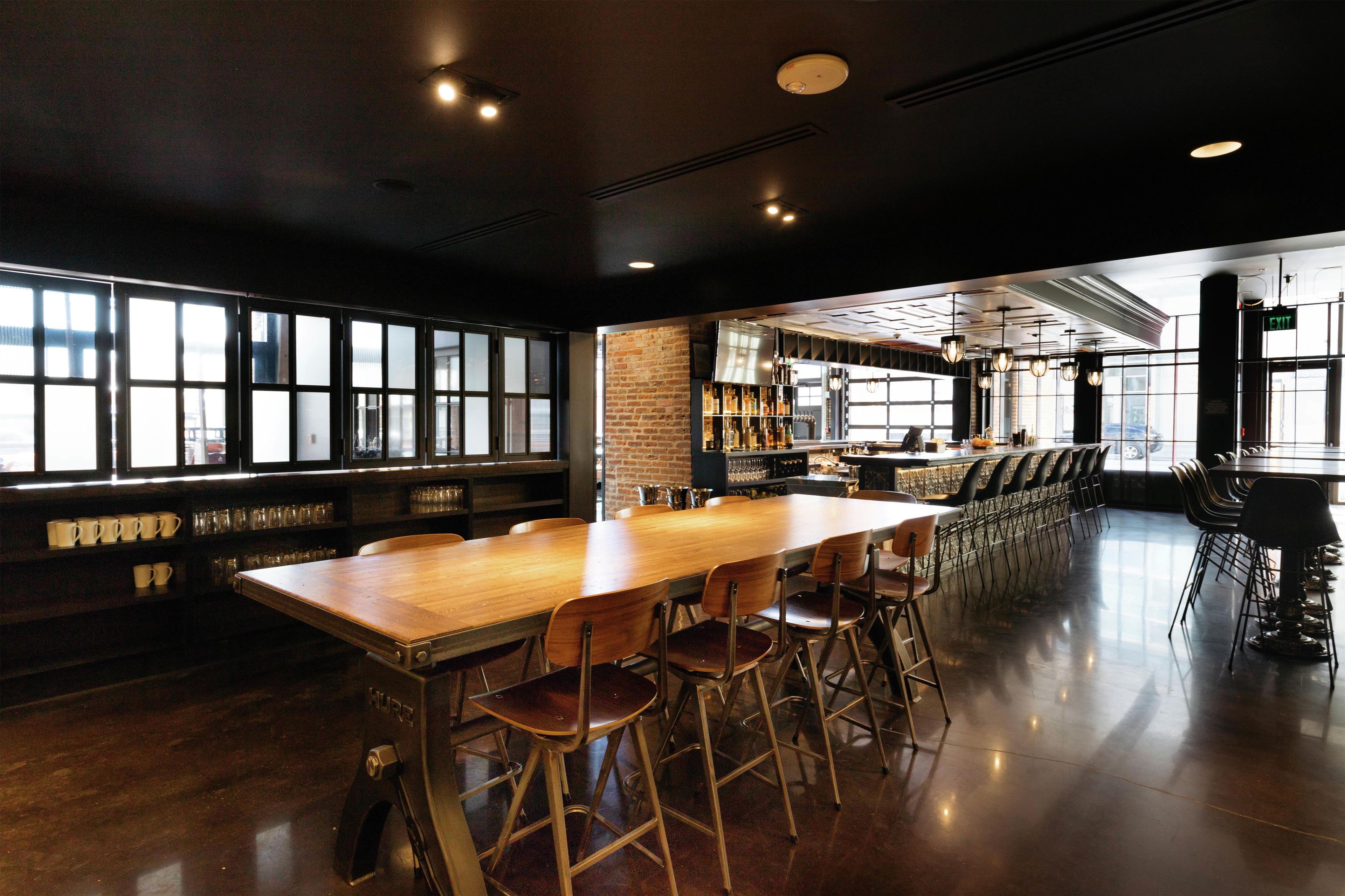 A spacious bar area with a long wooden table and multiple stools, a brick wall, and a bar counter with various bottles and glasses.
