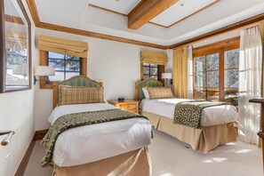 A hotel room with two beds, wooden beams on the ceiling, large windows with curtains, and a view of trees outside.