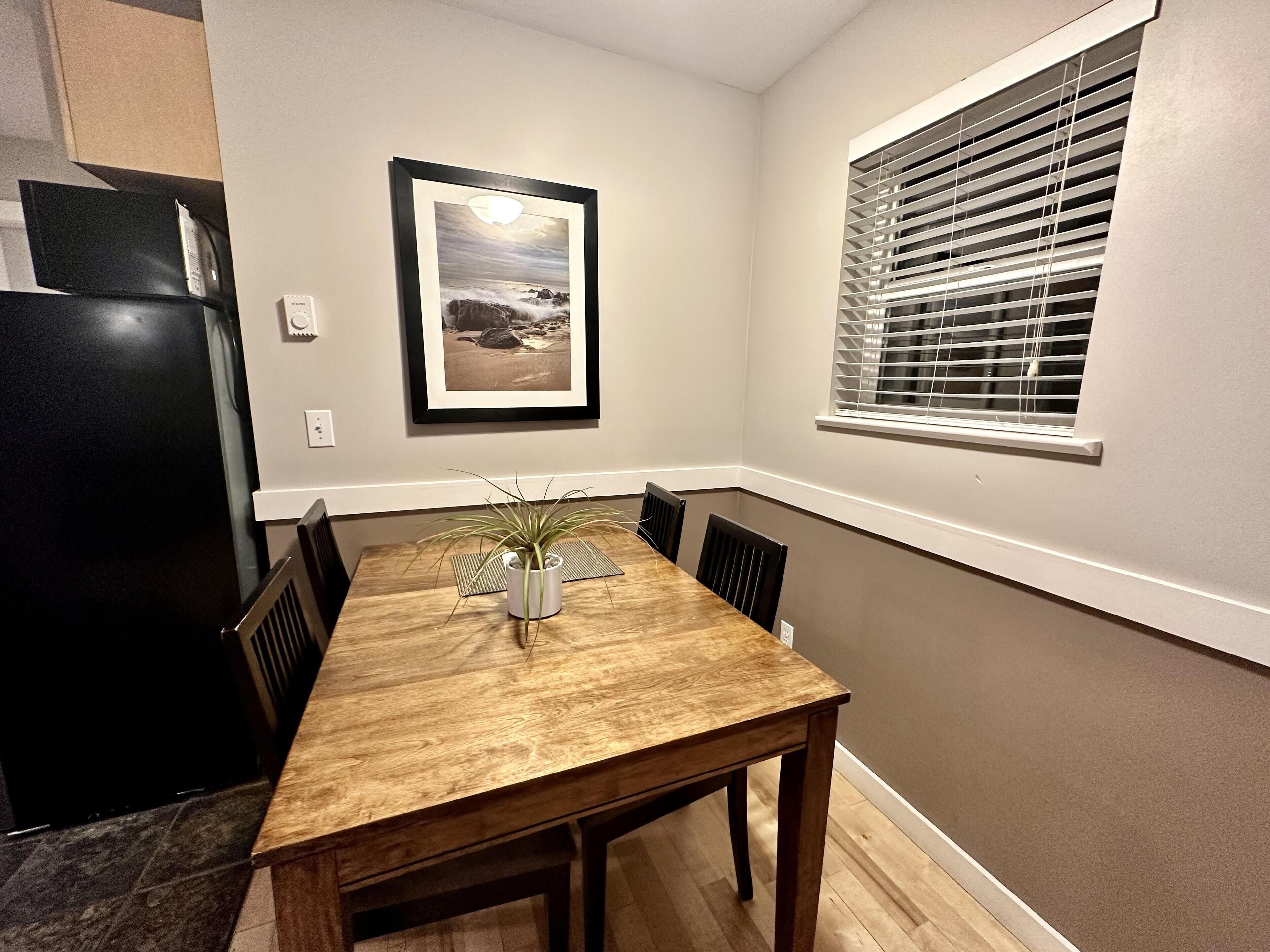 A dining area with a wooden table, chairs, a potted plant, a framed picture on the wall, a black refrigerator, and a window with blinds.