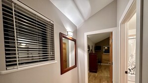 Un pasillo con piso de madera, una ventana con persianas, un espejo y una puerta que da a otra habitación.