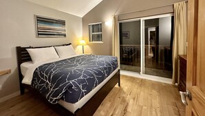 Un dormitorio con cama, mesita de noche, lámpara, ventana con persianas y puerta corrediza de vidrio que da a un balcón.