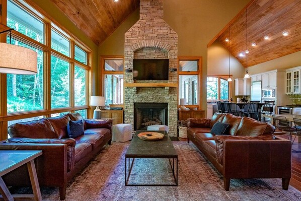 A spacious living room with leather sofas, a stone fireplace, and large windows offering a view of the outdoors.