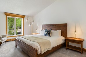Ein Schlafzimmer mit einem hölzernen Bett, einem Nachttisch, einem Sessel, einem Fenster mit Blick auf Bäume und einer Lampe.